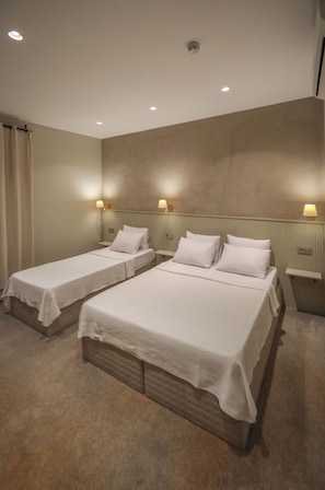 Habitación de hotel con dos camas individuales, una cama grande, una mesita de noche con lámpara y una luz en el techo.