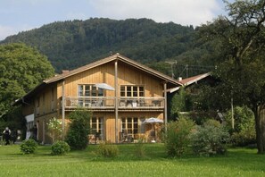 A two-story wooden house with a balcony, surrounded by a lush garden and a forested hill in the background.