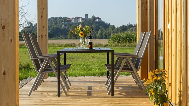 Una terraza de madera con una mesa y sillas, un florero con flores y una botella de vino.