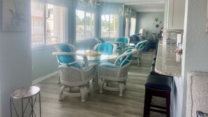 Une pièce spacieuse comprenant un coin repas avec des chaises en osier et une table ronde, de grandes fenêtres équipées de stores et un lustre.