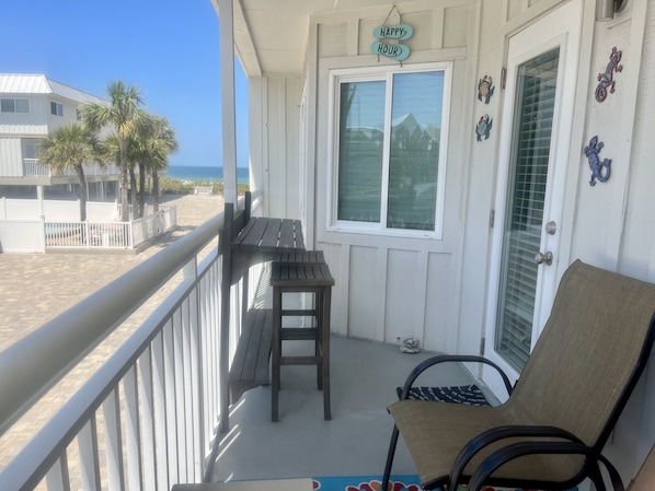 A balcony with a view of the beach, a wooden bench, a chair, and a 'Happy Hour' sign.