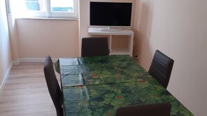 A dining area with a table covered in a green tablecloth with ladybug patterns, four chairs, a television on a white stand, and a window with blinds. - Piazza Roma holidays House  (Catania)