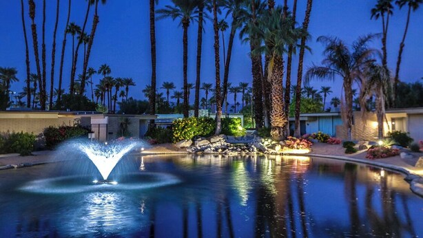 A nighttime view of a hotel pool with a water fountain, surrounded by palm trees and well-lit landscaping. - Mid Century Modern-Stunning Mountain Views, Quiet, Clean, Bright & Open Design. (Indian Wells)