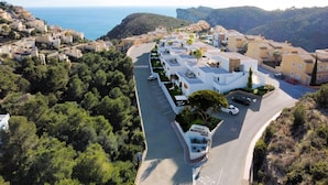 A coastal residential area with modern apartment buildings, a road with parked cars, and a sign indicating 'Pueblo Montecala'. - Apartamento Jardines PG018 - Benitachell, Costa Blanca (Benitachell)