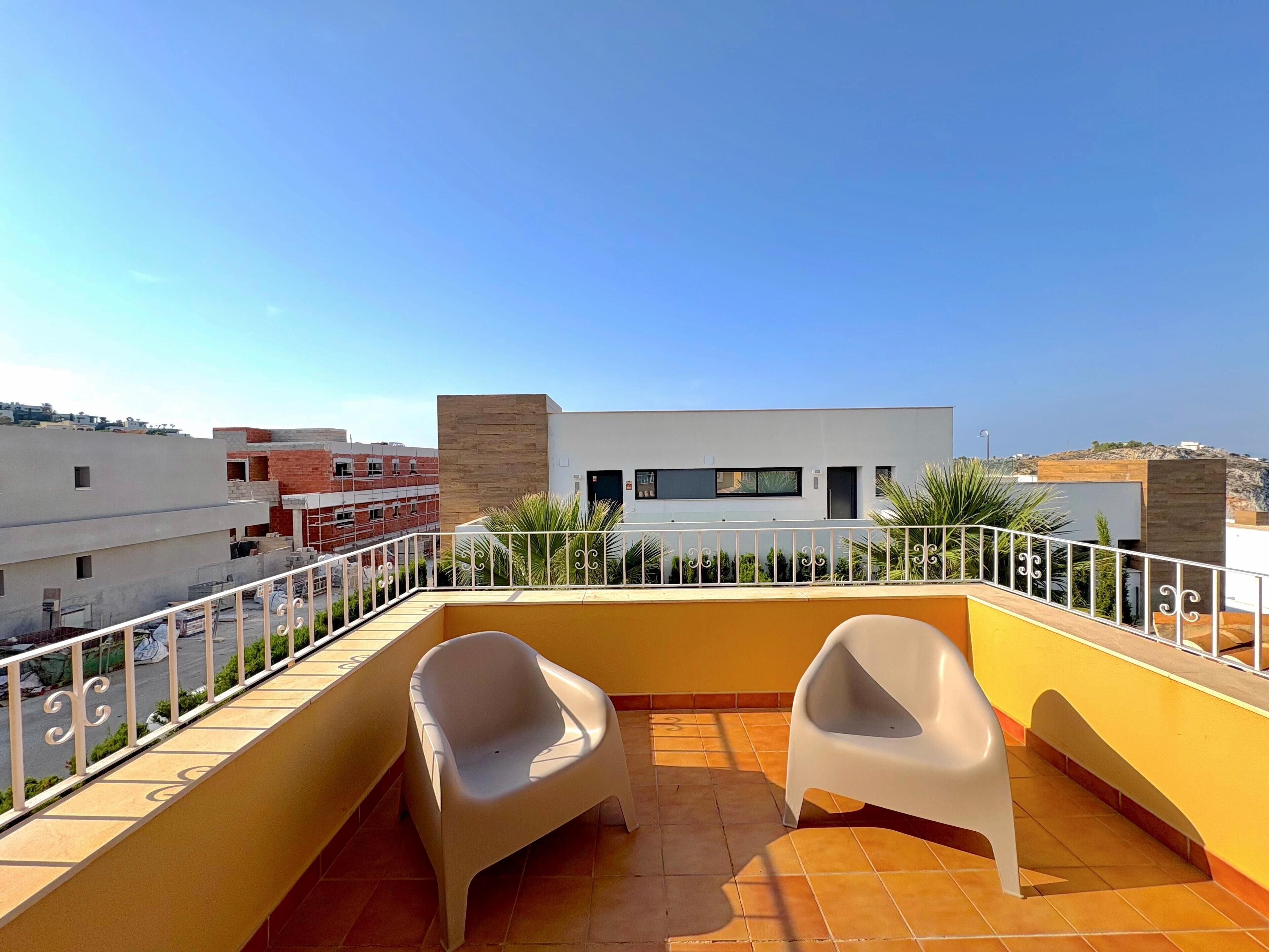 Ein Balkon mit zwei weißen Plastikstühlen, einem Fliesenboden und Blick auf moderne Gebäude im Hintergrund.