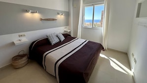 A bedroom with a bed, a bedside table, a wall-mounted shelf, a window with a view, and a hanging light fixture. - Apartamento Jardines PG018 - Benitachell, Costa Blanca (Benitachell)