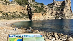 A rocky beach with a signboard displaying information about Cala Moraig. - Apartamento Jardines PG018 - Benitachell, Costa Blanca (Benitachell)