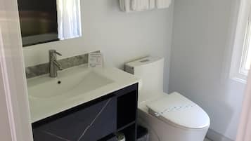 A modern bathroom with a white sink, mirror, and toilet. There are two towels hanging on a rack and a trash bin under the sink.