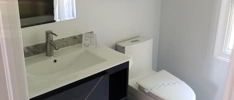 Une salle de bain moderne avec un lavabo, un miroir et des toilettes blancs. Deux serviettes sont suspendues à un porte-serviettes et une poubelle se trouve sous le lavabo.