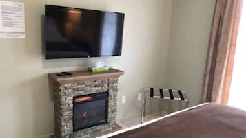 A room with a flat-screen TV mounted on the wall, a stone fireplace with a fire, a small table with a remote control, and a striped chair.