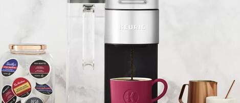 A Keurig coffee maker with a pink mug, a coffee pot, and a jar of coffee pods on a wooden surface.