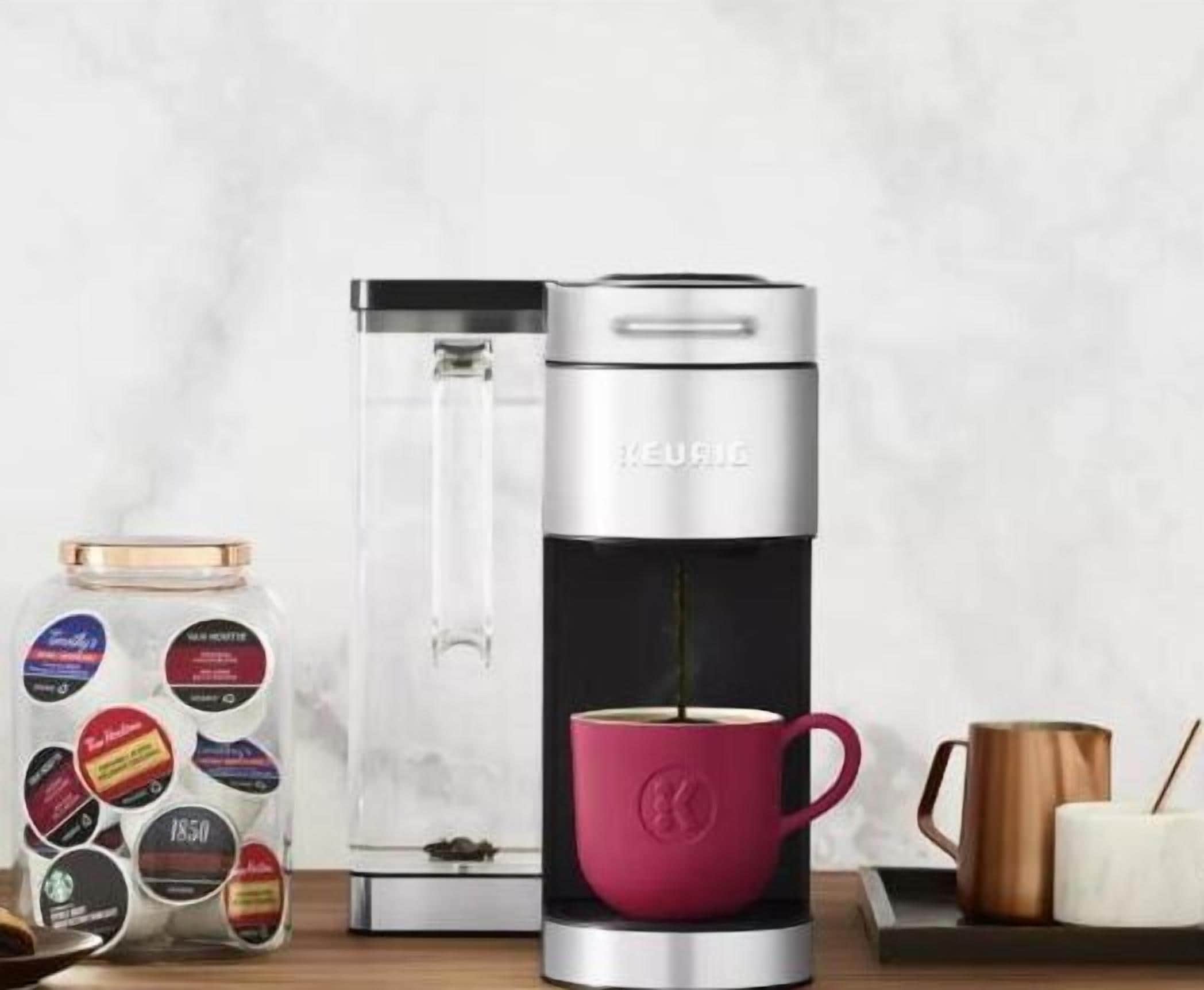 A coffee maker with a clear water reservoir, a red mug, and a jar of coffee pods.