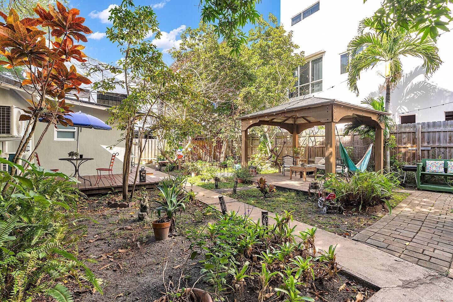 Un patio couvert avec des sièges d’extérieur, entouré d’une végétation luxuriante et avec un bâtiment blanc en arrière-plan.