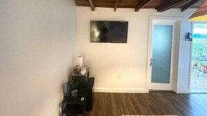 A room with a wooden ceiling, a flat-screen TV mounted on the wall, a door leading to an outdoor area, and a small table with a lamp. - 1920s Bungalow Studio w/ Courtyard Oasis (Miami)
