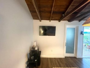 Une pièce avec un plafond en bois, un téléviseur à écran plat fixé au mur, une porte donnant sur un espace extérieur et une petite table avec une lampe.