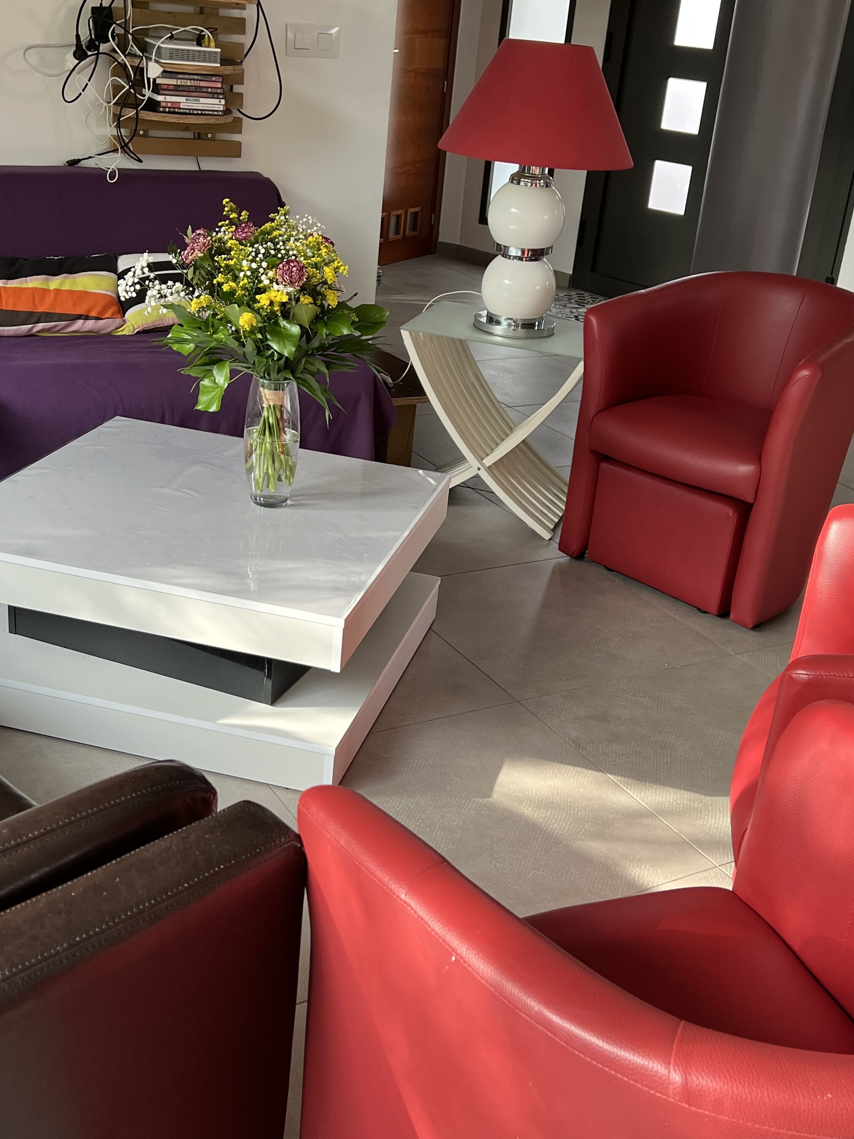 Un salon moderne avec un canapé, un fauteuil rouge, une table basse ornée d’un vase de fleurs et une lampe.