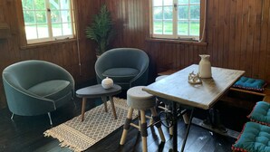 Una habitación acogedora con paredes de madera, dos ventanas adornadas con cortinas a rayas, una planta en maceta, una mesa con una jarra y dos sillas.