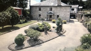 A stone building with a central entrance, surrounded by a gravel driveway and landscaped garden with various shrubs and a small tree.