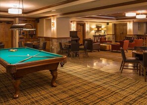 Una mesa de billar en una habitación con alfombra estampada, varias sillas y una zona de bar con televisor.