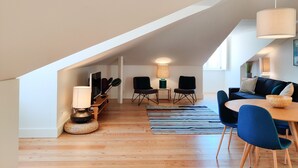 Un loft moderne avec un plafond haut, un plancher en bois, un coin repas avec une table ronde et des chaises bleues, et un espace de vie avec un canapé foncé et des chaises noires.