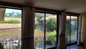 A room with large windows, a balcony, and a view of a garden and buildings.