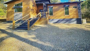 A two-story log cabin with a stone foundation, a wooden staircase, and a gravel driveway.