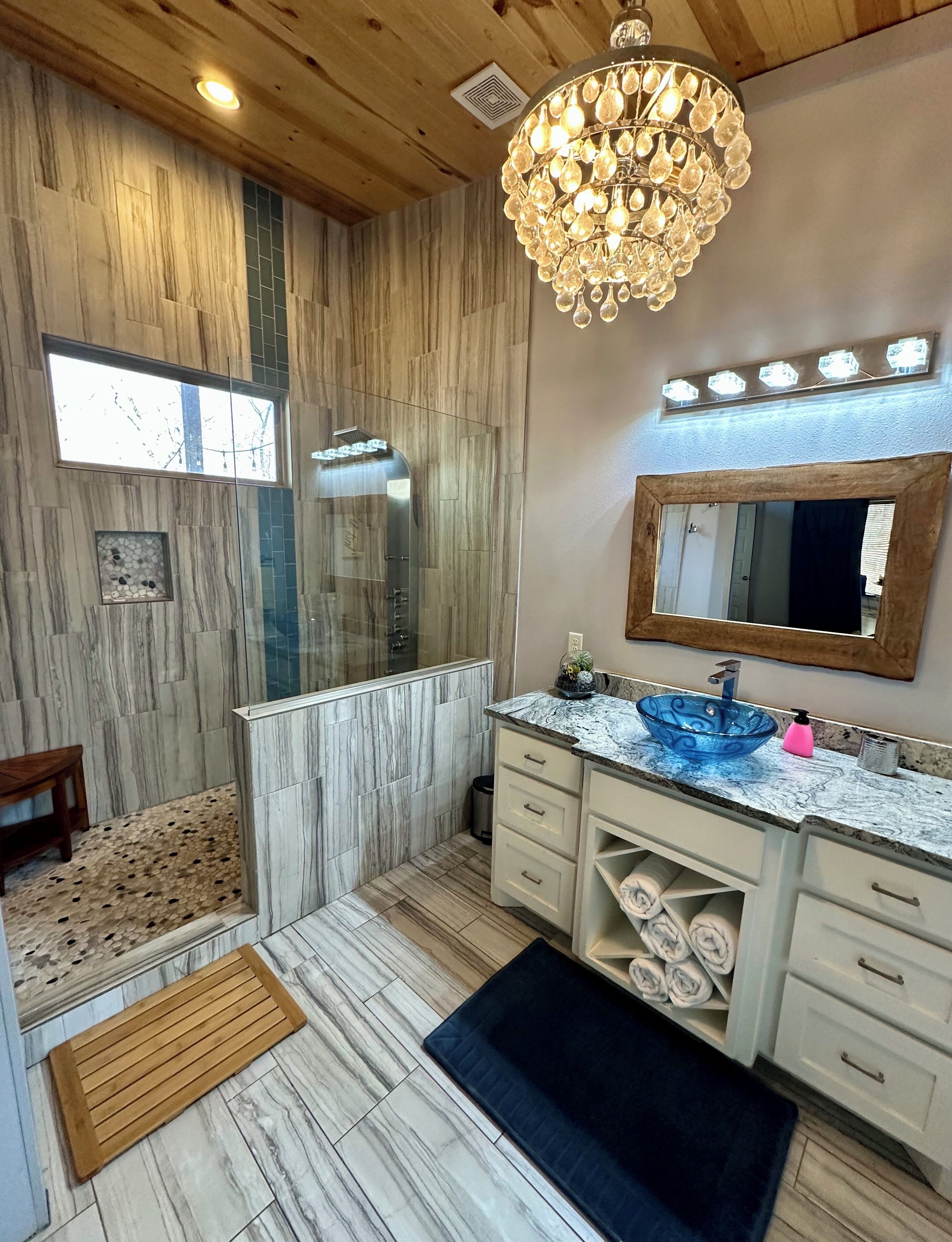 A modern bathroom with a marble floor, a glass shower enclosure, a vanity with a blue glass bowl sink, and a large mirror above the sink.