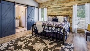 A bedroom with a wooden ceiling, a bed with patterned bedding, a ceiling fan, and a large window with blinds.