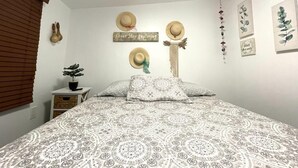 A bedroom with a patterned bedspread, a small bedside table with a plant, and wall decorations including hats and a sign.