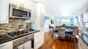 A modern kitchen with stainless steel appliances, a dining area with a wooden table and wicker chairs, and a view of the living room in the background. - Beach across the street, Alamitos Bay steps away! No hidden Fees! (Long Beach)