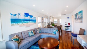 A modern living room with a sectional sofa, a round coffee table, and artwork depicting a beach scene. - Beach across the street, Alamitos Bay steps away! No hidden Fees! (Long Beach)