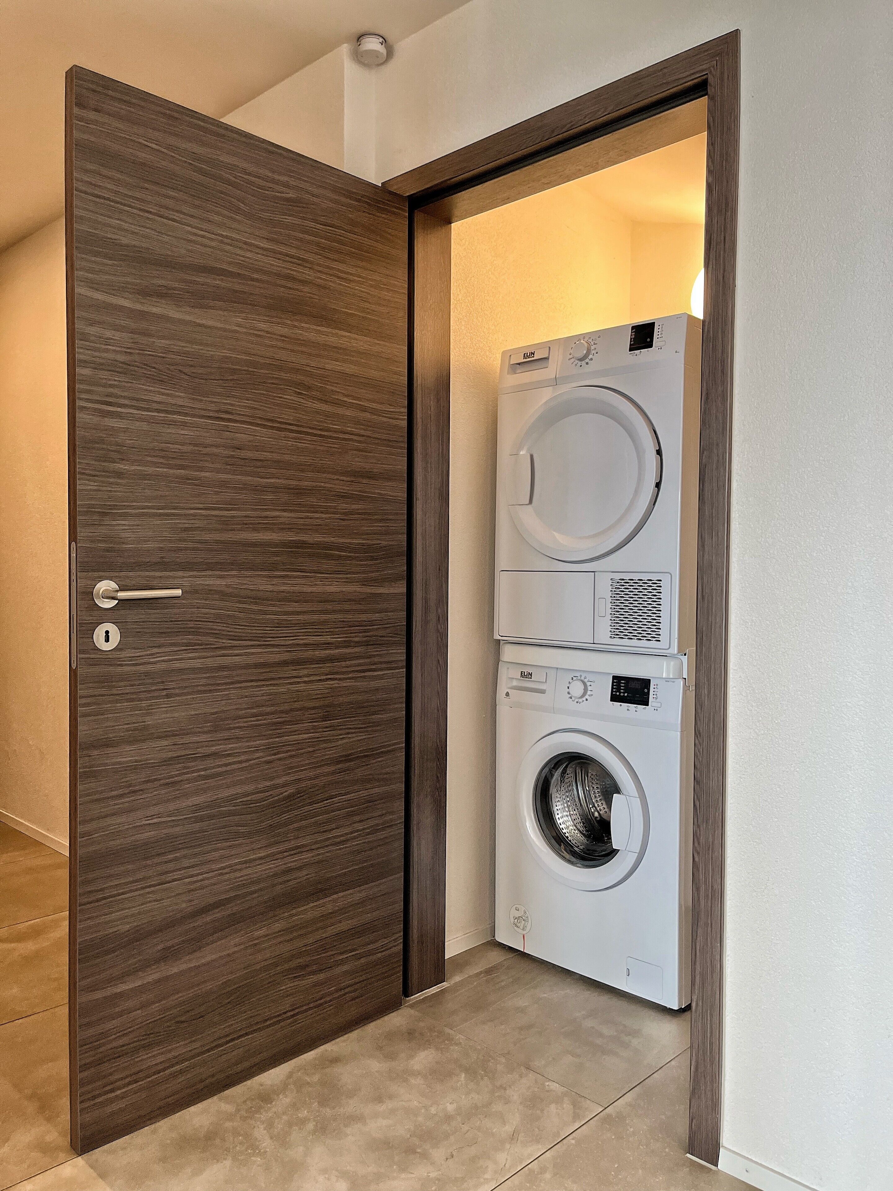 Ein Waschraum mit einer Frontlader-Waschmaschine und einem Trockner, einer Holztür und einem gefliesten Boden.