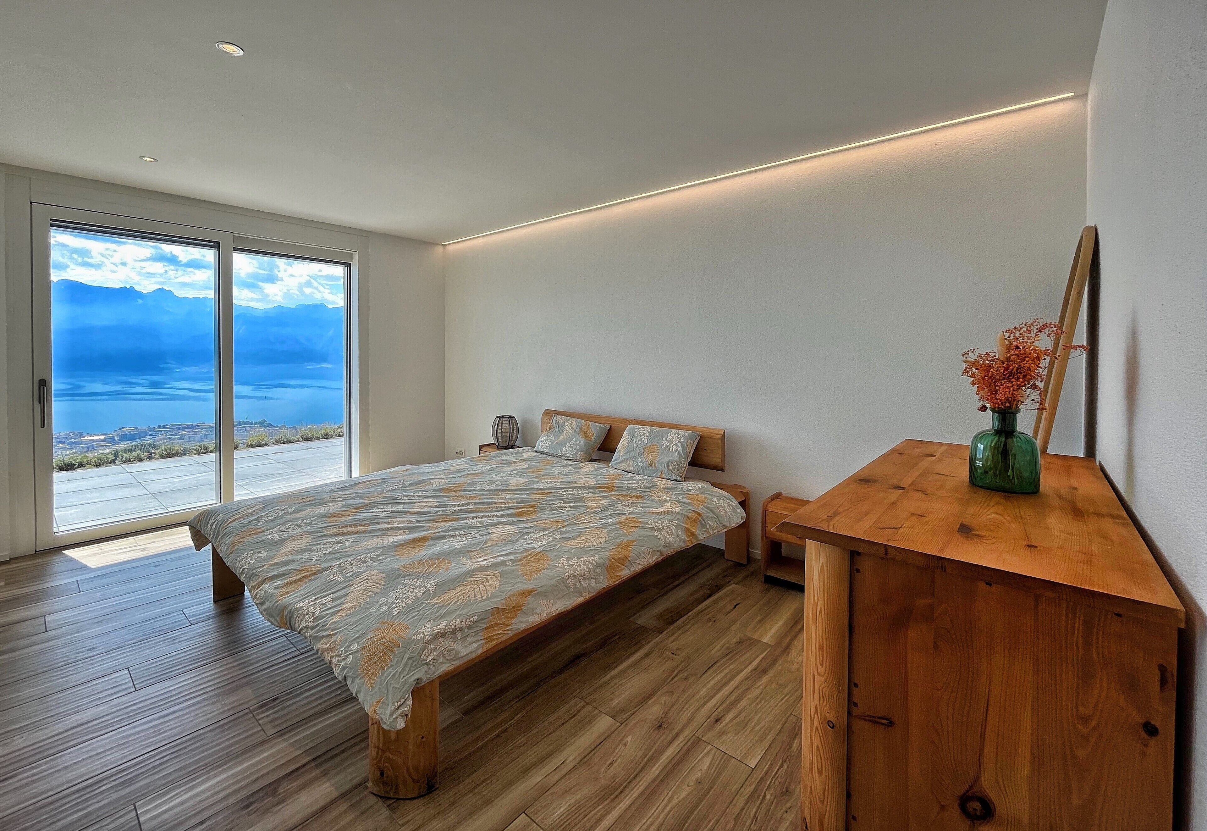 A bedroom with a large bed, wooden flooring, a wooden nightstand, a mirror, and a view of the outdoors.