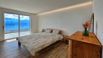 A bedroom with a large bed, wooden flooring, a wooden nightstand, a mirror, and a view of the outdoors.