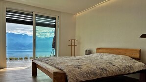 Ein Schlafzimmer mit einem hölzernen Bett, Blick auf Berge und Wasser durch ein großes Fenster sowie einer Stehlampe.