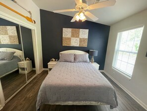 A bedroom with a bed, a ceiling fan, a nightstand, a lamp, a mirror, and a window with blinds.