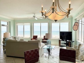 A living room with a sofa, chairs, a TV, a chandelier, and large windows overlooking the ocean.