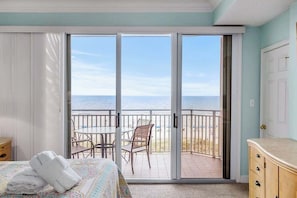 A room with a bed, a set of white towels, a table with chairs, and a view of the beach through a sliding glass door.