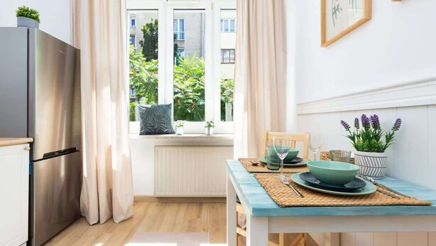 A compact kitchen with a modern refrigerator, a window with a view of greenery and buildings, a dining table with a blue tablecloth, and a potted plant on the windowsill. - Stylish Studio in Mokotow | Close to Metro and Parks (Warszawa)