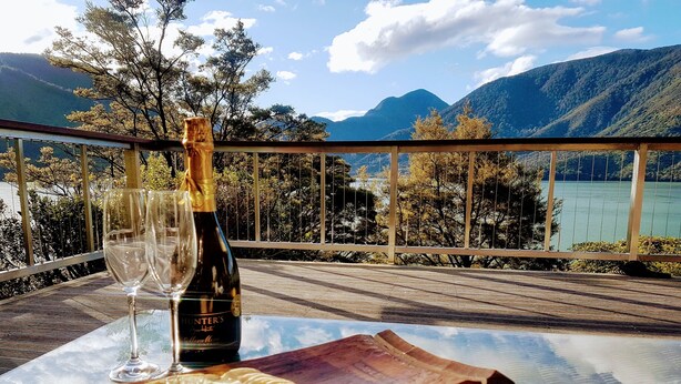 A table with a bottle of Hunter's 2018 Chardonnay, a glass, and a cheese wheel on a wooden board, overlooking a lake and mountains. - 🌿 Birdsong Hideaway 🌿 (Moetapu Bay)