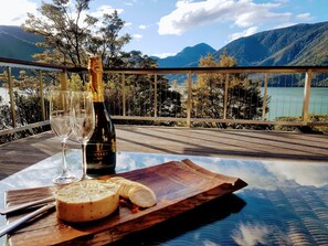 Une table sur laquelle se trouvent une bouteille de Chardonnay Hunter, millésime 2018, un verre et une portion de fromage, le tout posé sur une planche en bois, avec une vue sur un lac et des montagnes.