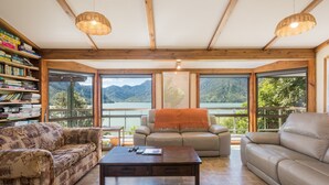 A spacious living room with a large window overlooking a lake and mountains, furnished with a sofa, armchair, and coffee table. - 🌿 Birdsong Hideaway 🌿 (Moetapu Bay)