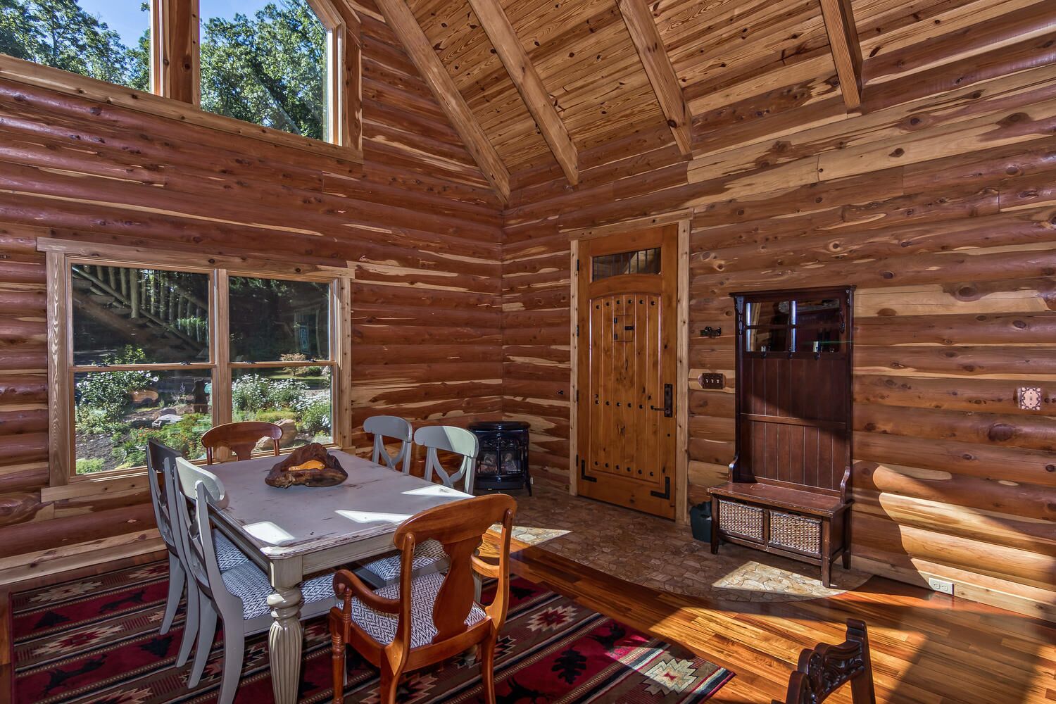 A log cabin interior with a dining table, chairs, a fireplace, and a large window offering a view of the outdoors.