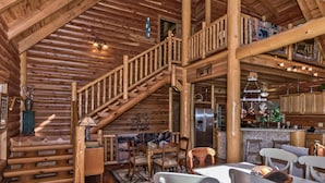 Interior de una cabaña de troncos con escalera de madera, comedor, cocina y habitación en el altillo.