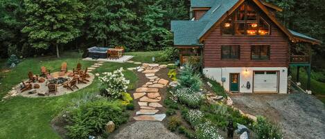 A log cabin with a stone pathway leading to a hot tub area surrounded by greenery.