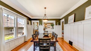 A dining area with a wooden table, chairs, and a chandelier. There are windows on one side and a white wall on the other. - The Proctor House: North End, Old Town, Ruston (Tacoma)