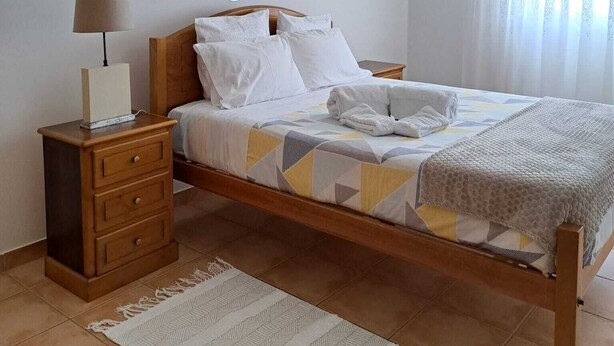 A bedroom with a wooden bed, white bedding, a bedside table with a lamp, a wall-mounted mirror, and a patterned rug. - Violante Apartment Beach (Foz do Arelho)