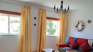 A living room with a sofa, a coffee table, and a chandelier. There are windows with curtains and a view outside. - Violante Apartment Beach (Foz do Arelho)