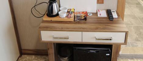 A compact hotel room with a television mounted on a wooden cabinet, a small refrigerator, a waste bin, and a coffee maker.