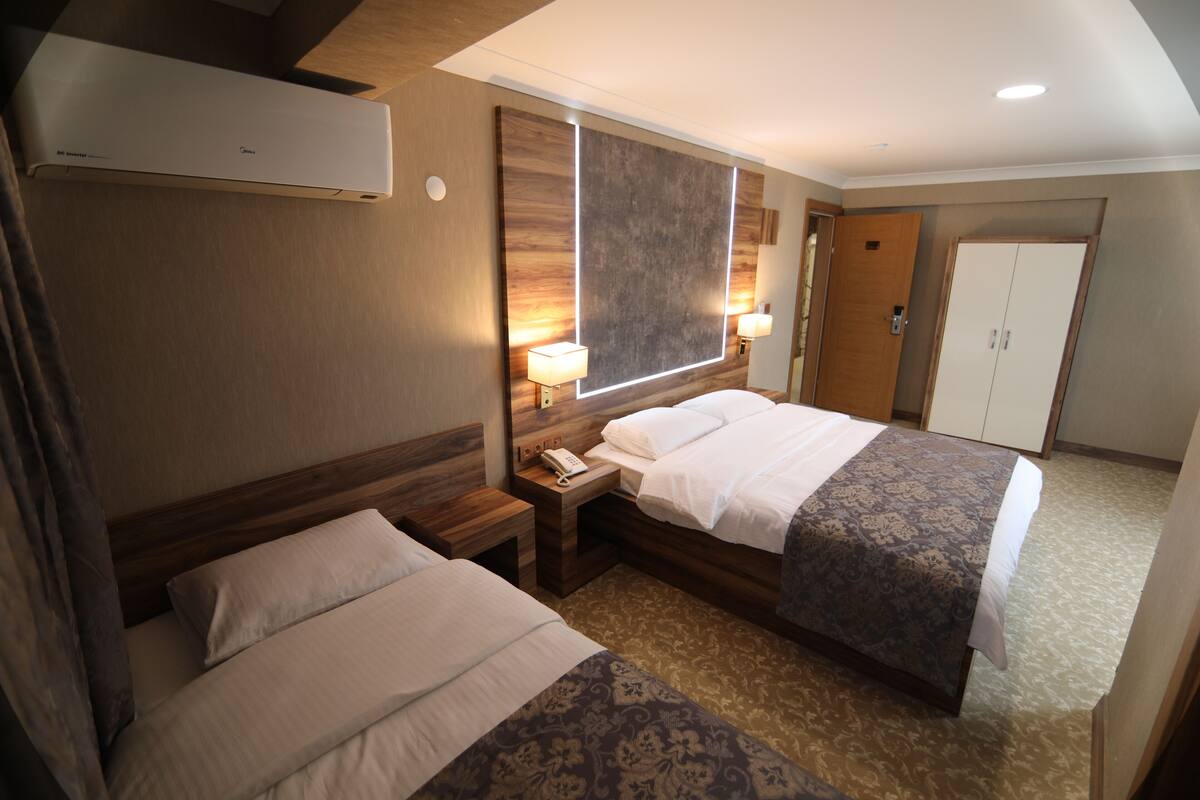 A hotel room with two beds, a large mirror, a bedside table with a lamp, and a wardrobe.